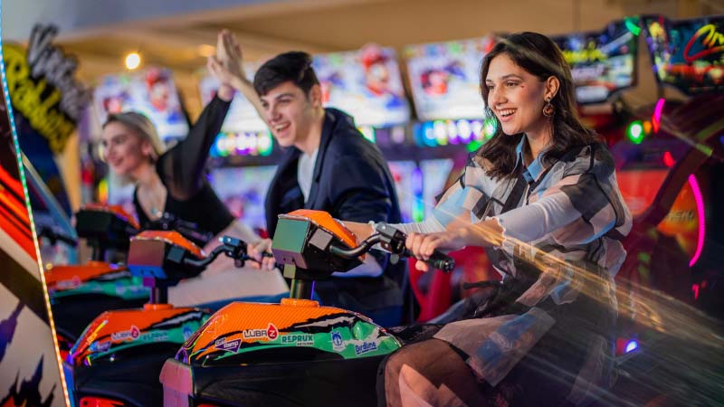 Three people joyfully ride arcade motorcycles, surrounded by vibrant lights and screens. The scene exudes excitement and fun in a lively arcade setting.