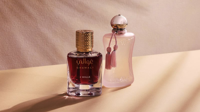 Two elegant perfume bottles on a soft beige surface. The left bottle is dark with a gold cap labeled "Ghawali." The right is pink, ornate, and tasseled.